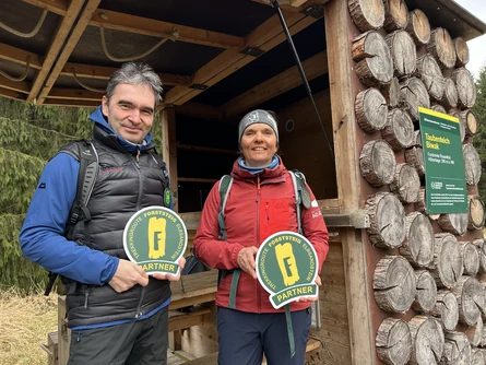 Ein Mann und eine Frau in Wanderbekleidung stehen in einer Hütte und halten jeweils ein Schild "Forststeig-Partner" in den Händen. An der Hütte hängt ein Schild mit der Aufschrift "Taubenteich-Biwak".