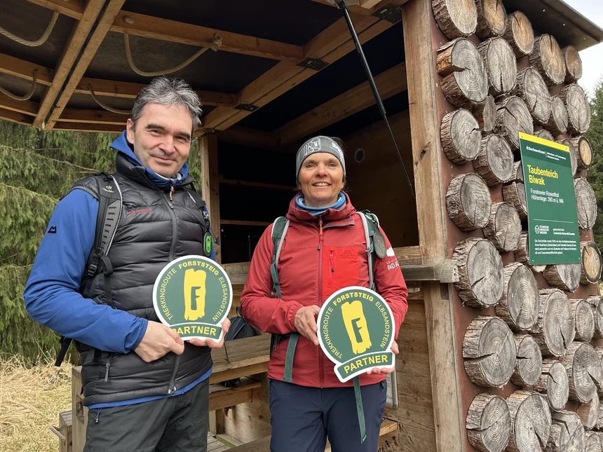 Ein Mann und eine Frau in Wanderbekleidung stehen in einer Hütte und halten jeweils ein Schild "Forststeig-Partner" in den Händen. An der Hütte hängt ein Schild mit der Aufschrift "Taubenteich-Biwak".