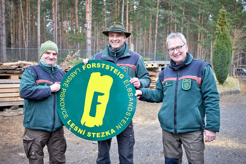Die Hüttenwarte Wolfram Claus (links) und Jens Dzikowski (rechts) sind auch wieder auf dem Forststeig unterwegs. 3 Männer in Forstbekleidung und großem Schild mit Forststeiglogo