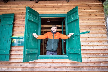 junger Mann &ouml;ffnet die Fensterl&auml;den an einer Forsth&uuml;tte