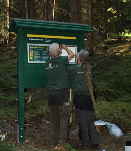 Zwei Mitarbeiter von Sachsenforst schrauben Schilder an eine gro&szlig;e Tafel