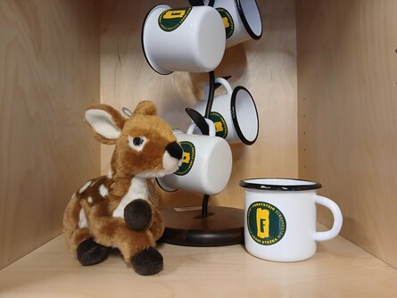 Ein Plüschreh liegt neben Emaille-Tassen mit Forststeig-Logo.