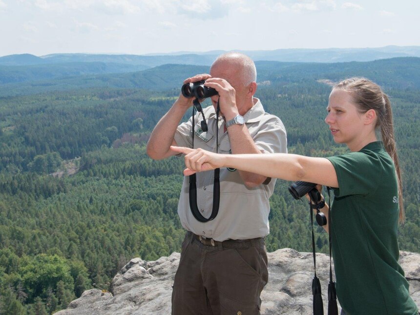 Ein Mitarbeiter und eine Mitarbeiterin der Naturwacht stehen auf Felsen und beobachten mit Ferngläsern Aktivitäten in der Landschaft